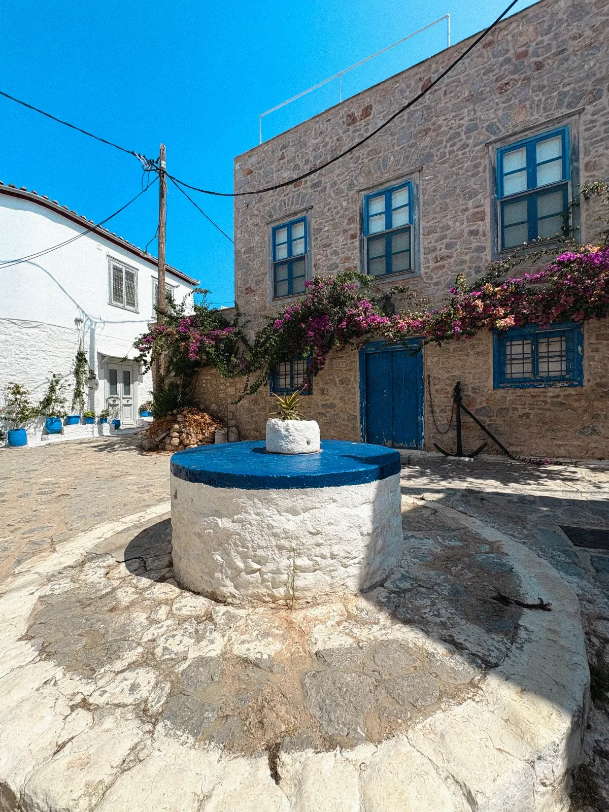 Weiß getünchter Brunnen in einem griechischen Innenhof neben einem Steinhaus mit blauen Fensterläden